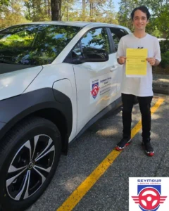 A young man in a white T-shirt, black pants, and black sneakers with red soles holds a yellow form beside a white Seymour Driving School car.