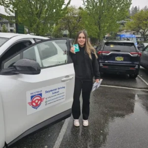A young woman in a black hoodie and black pants holds up a green N sign beside the open door of a white Seymour Driving School car on wet pavement.