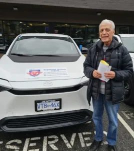 An older man in a black puffer jacket and blue jeans holds yellow and white papers beside a white Seymour Driving School car.