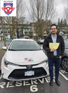 A graduate in a black jacket holds a yellow form beside a white Seymour Driving School car.