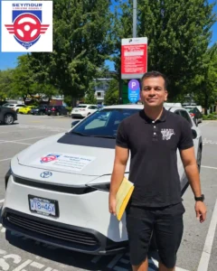 A graduate in a black polo shirt holds a yellow folder beside a white Seymour Driving School car.