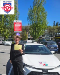 A woman in a black shirt and sunglasses holds a yellow form beside a white Seymour Driving School car.